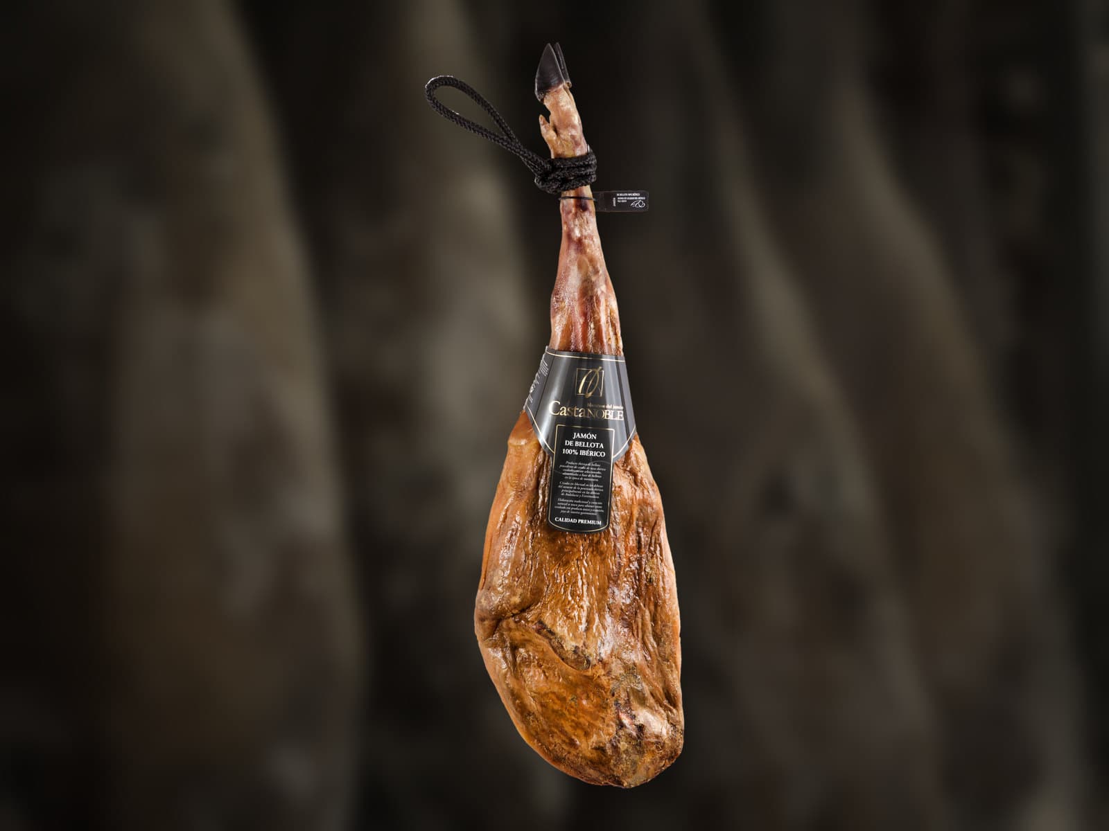 Un jamón de bellota 100 % ibérico de Castanoble, entero y con su precinto negro, colgado ante los jamones de nuestro secadero en penumbra.
