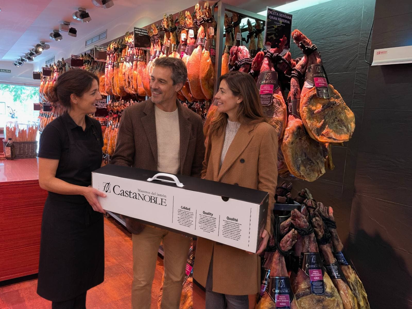 Recreación: una pareja entrega una caja de Castanoble a una dependienta en la tienda de Barcelona, ante la pared de jamones